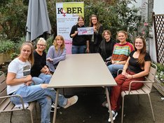 6 Personen sitzen um einen beigen Tisch und gucken in die Kamera. Sonja lemke hält einen symbolischen Scheck mit der Leiterin von Kober Tamara Degenhardt-Kirchmeyer in der Hand
