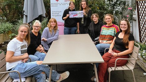6 Personen sitzen um einen beigen Tisch und gucken in die Kamera. Sonja lemke hält einen symbolischen Scheck mit der Leiterin von Kober Tamara Degenhardt-Kirchmeyer in der Hand