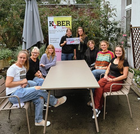 6 Personen sitzen um einen beigen Tisch und gucken in die Kamera. Sonja lemke hält einen symbolischen Scheck mit der Leiterin von Kober Tamara Degenhardt-Kirchmeyer in der Hand