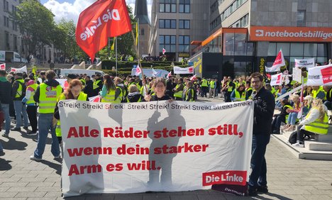 Menschen halten vor eine verdi-Demo in gelben Warnwesten ein Linke Transparent