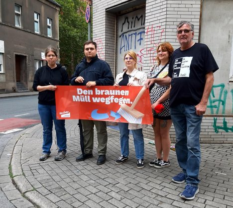Fünf Politiker:innen stehen auf einer Straße und halten ein Banner hoch auf welchem steht: Den braunen Müll wegfegen.