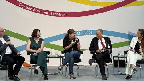 5 Personen sitzen auf einer Bühne. Sonja Lemke in der Mitte und redet in das Mikrofon. Auf der Wand hinter der Bühne steht Tag der Ein- und Ausblicke